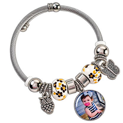 Angled view of A Memory to Wear photo bracelet with a round child portrait charm, floral beads, and silver-tone owl and butterfly charms.