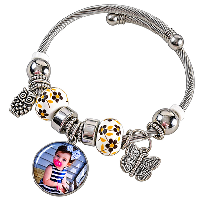 A Memory to Wear bracelet with a blank round photo setting, silver-tone cable band, yellow floral beads, and butterfly and owl charms.