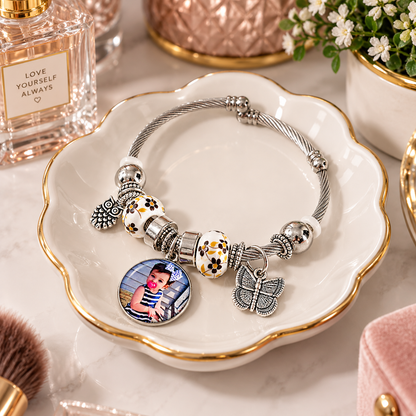 A Memory to Wear personalized photo bracelet resting in a white, gold-rimmed jewelry dish on a dresser beside flowers and perfume.