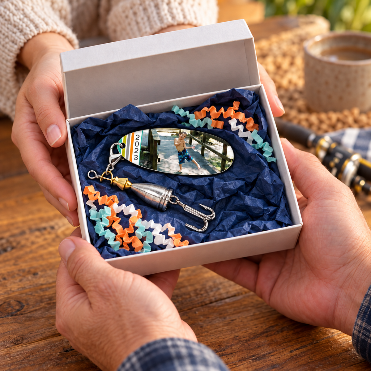 Hands presenting a personalized photo fishing lure in an open white gift box with navy tissue and orange, teal, and white crinkle paper.