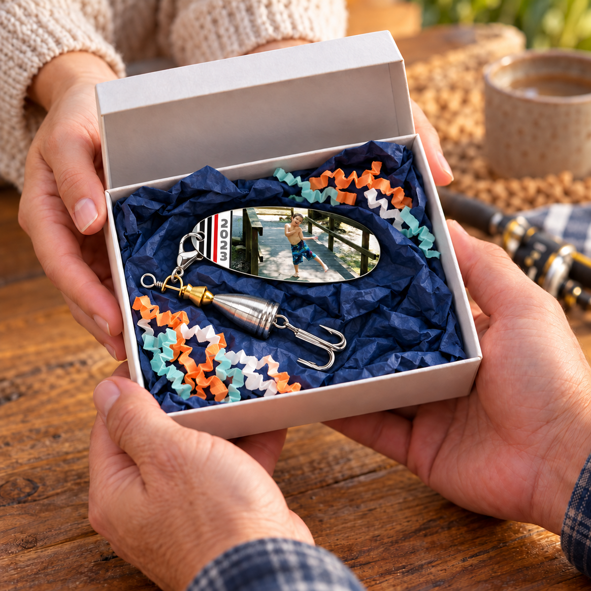 Hands presenting a Galactic Dad photo fishing lure in an open white gift box with navy tissue and orange, teal, and white crinkle paper.