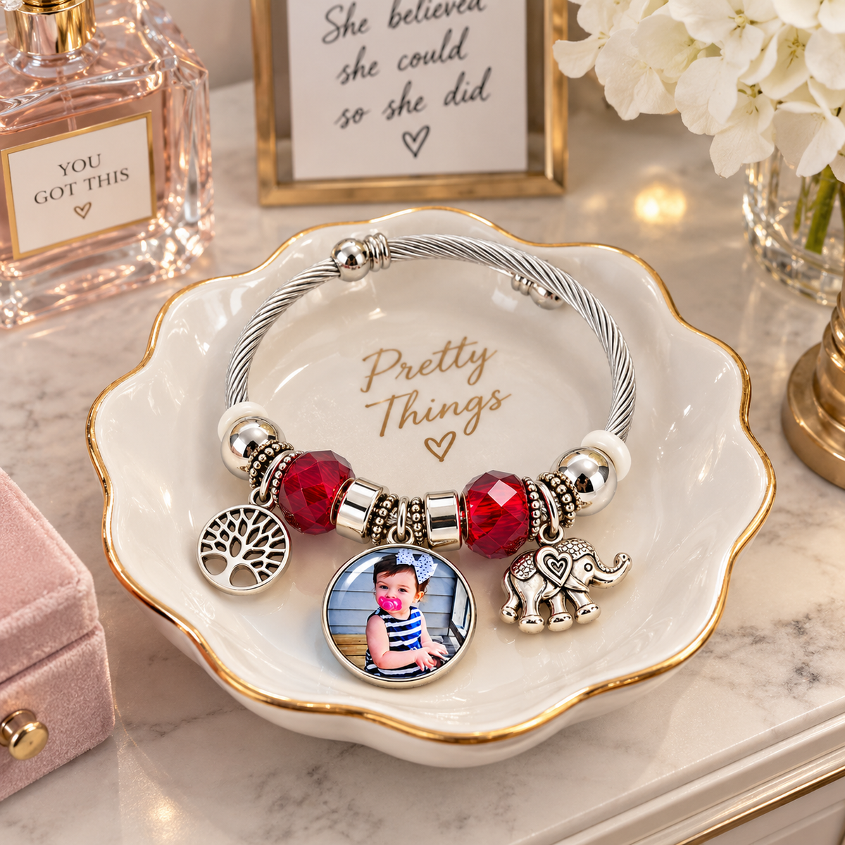 Forever in My Heart personalized bracelet resting in a white, gold-rimmed jewelry dish beside perfume and white flowers.