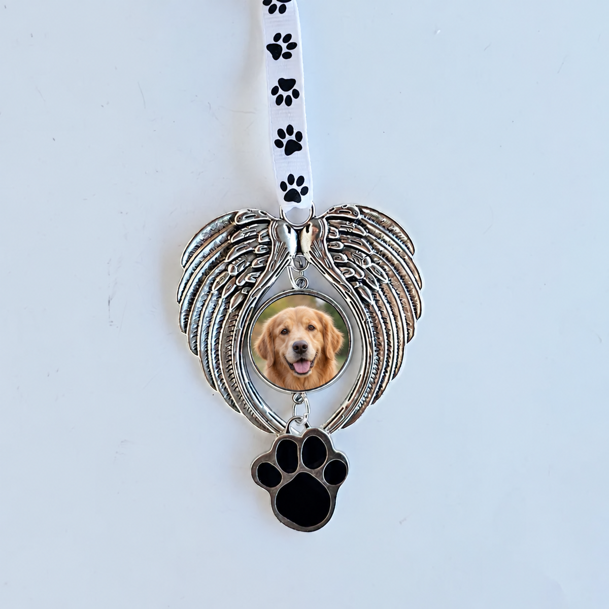 Silver-tone angel wing pet memorial ornament with a golden retriever photo, black paw-print charm, and white paw-print ribbon against a pale background.