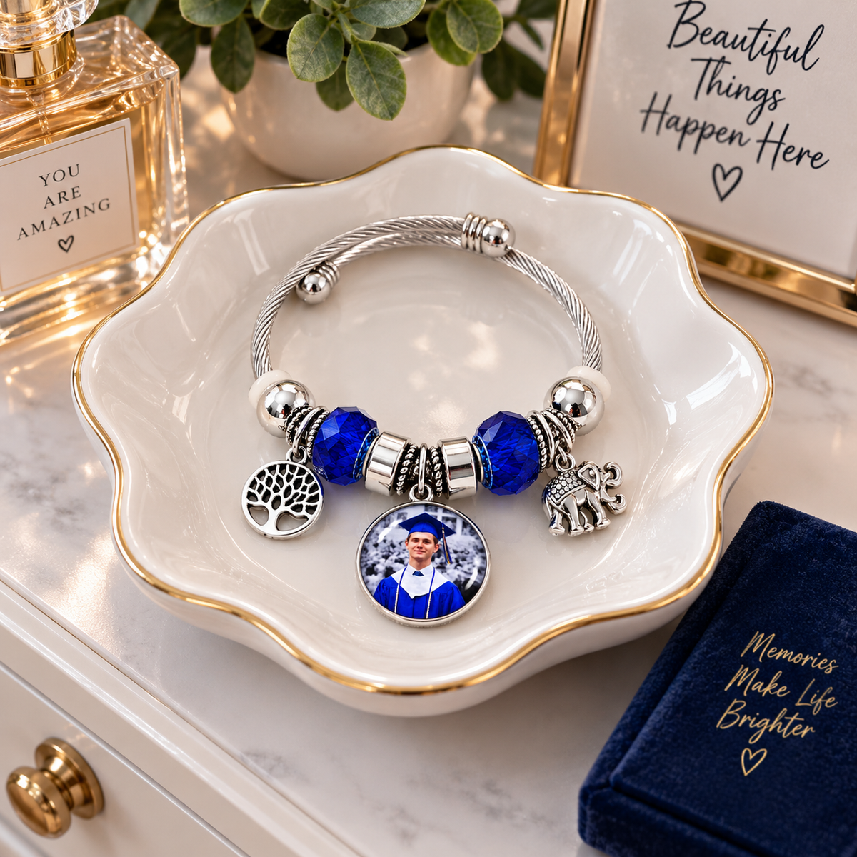 Rooted in Memories personalized photo bracelet resting in a white, gold-rimmed jewelry dish beside perfume, greenery, and a navy notebook.