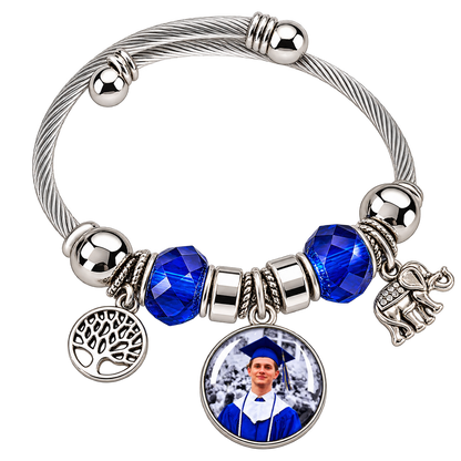 Angled view of Rooted in Memories personalized graduation photo bracelet with blue beads, a silver-tone cable band, and dangling tree and elephant charms.