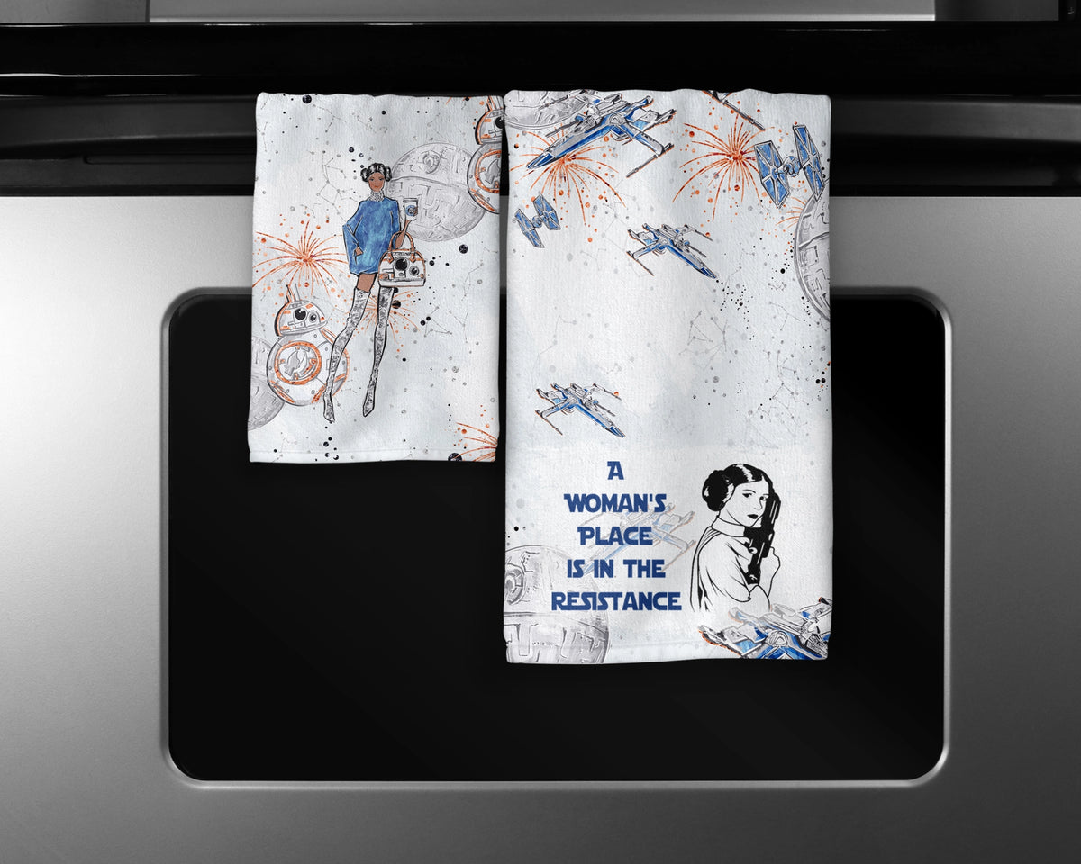 A Woman’s Place Is In The Resistance two-piece hand towel set featuring space-inspired characters and blue Resistance statement lettering, hanging side by side on oven handle